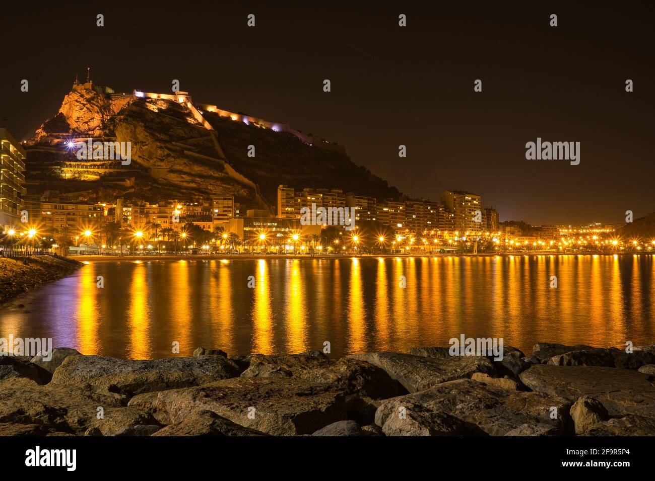 Strand am Fuße eines Schlosses mit Reflexionen der Lichter der Stadt im Meer, in Spanien Stockfoto