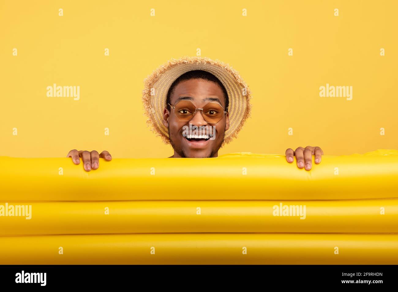 Millennial Black Guy in Strohhut und Sonnenbrille versteckt sich dahinter Aufblasbare Lilo auf gelbem Studiohintergrund Stockfoto