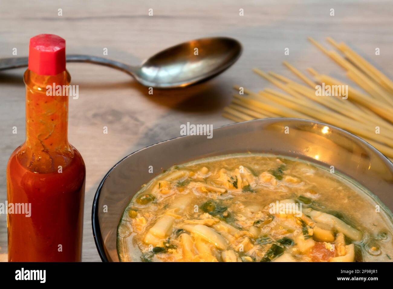 Würzige asiatische Nudelsuppe in einer Glasschüssel, Tabasco-Sauce und ungekochte Nudel auf einem Holzbrett serviert Stockfoto