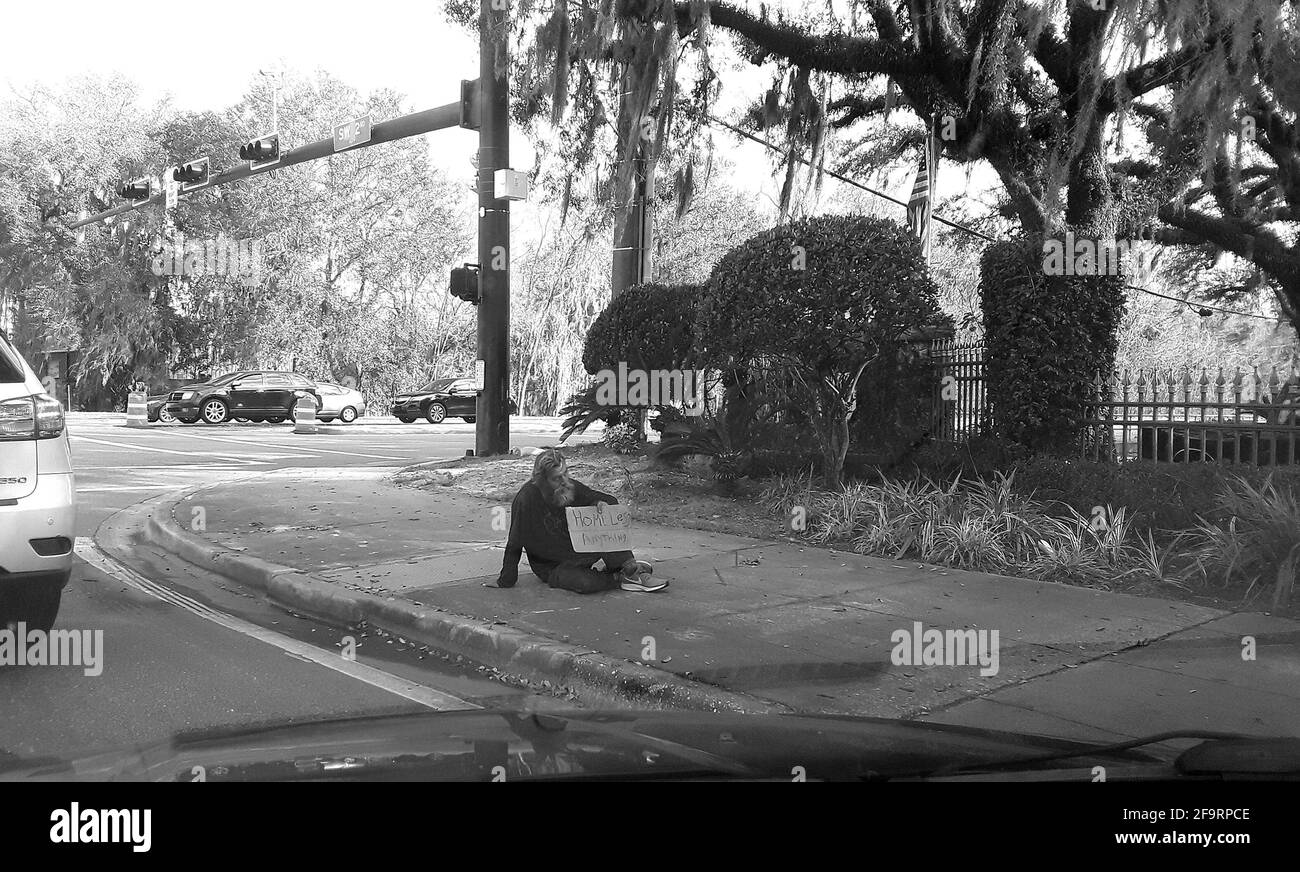 Obdachloser, der auf dem Bürgersteig sitzt und ein Schild in Gainesville, Florida hält. Stockfoto