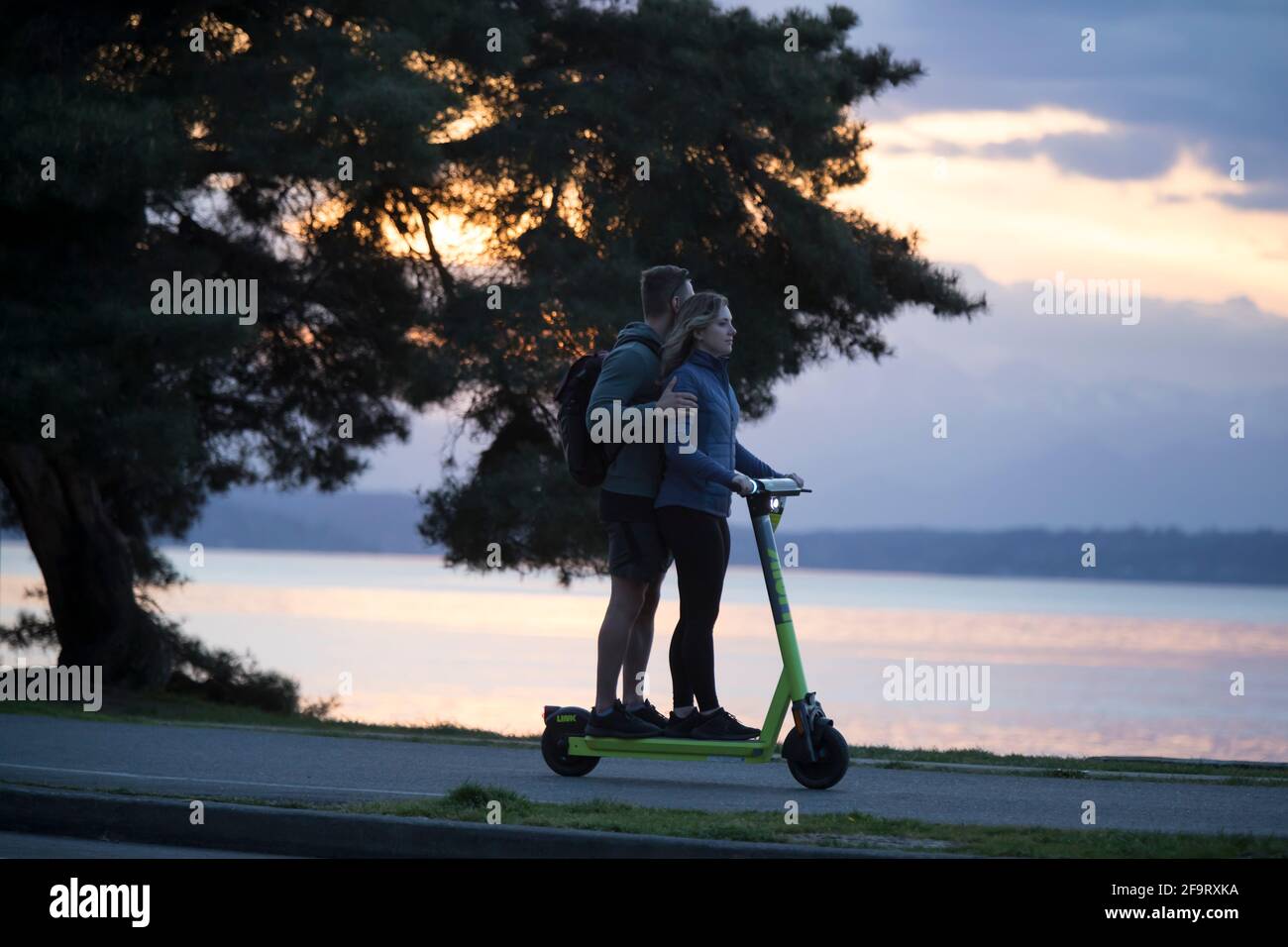 Ein junges Paar teilt sich am Montag, den 19. April 2021, einen Roller auf dem Alki Trail, während die Sonne über den Olympic Mountains in Seattle untergeht. Stockfoto