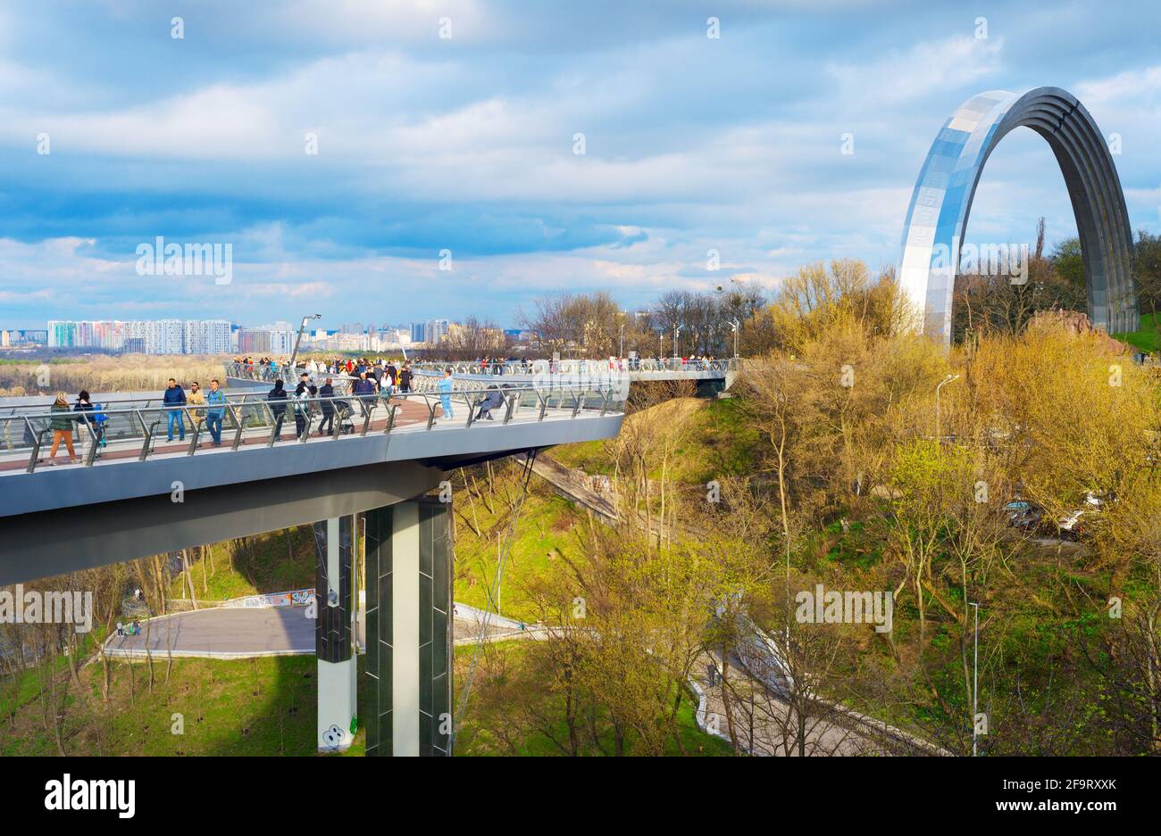 KIEW, UKRAINE - 18. APRIL 2021: Menschen gehen an der neuen Fußgängerbrücke mit dem Fahrrad, im Hintergrund der Völkerfreundschaftsbogen Stockfoto