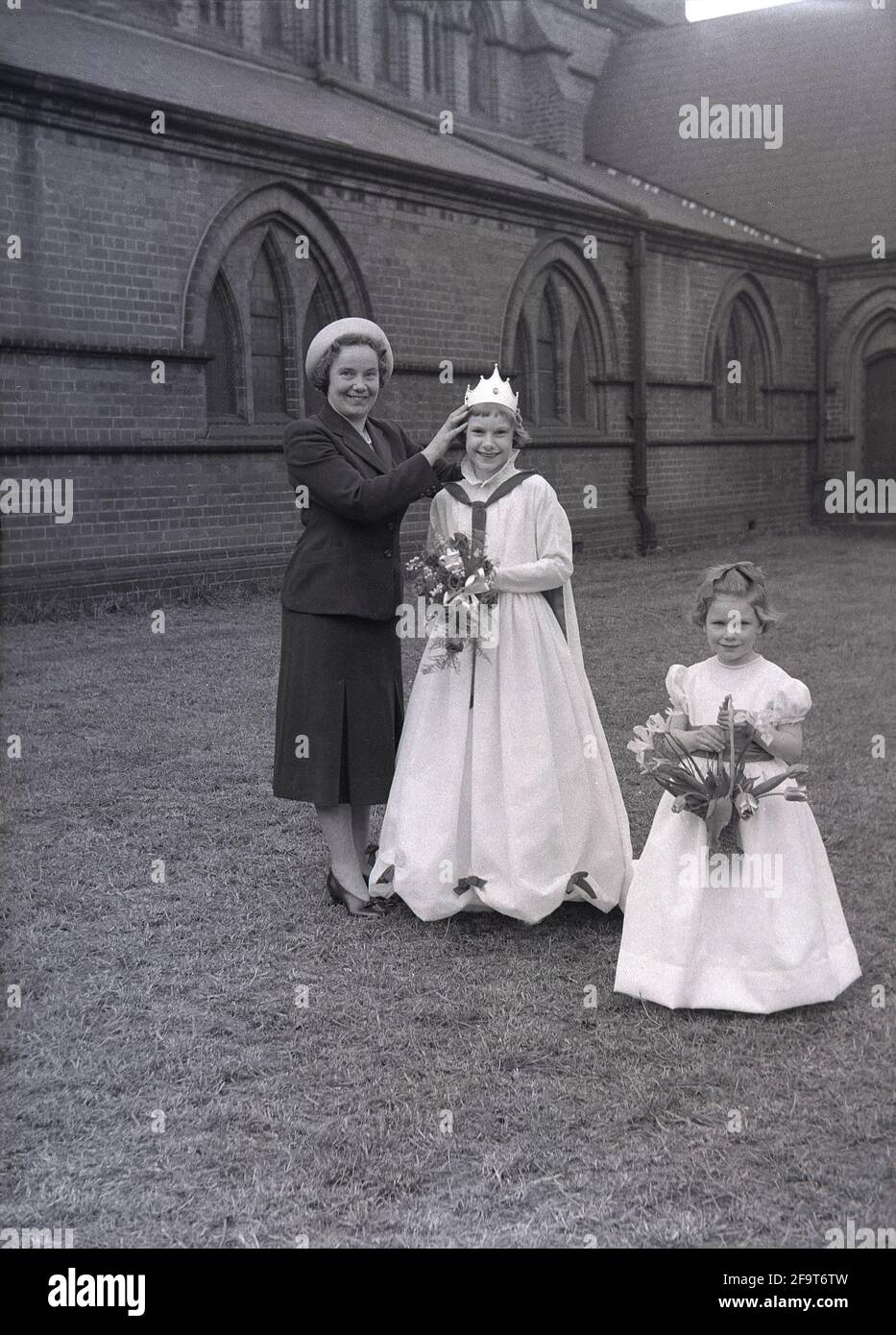 1956, draußen auf dem Gelände einer Kirche, eine stolze Mutter mit ihrer süßen jungen Tochter, die ihre May Queen-Krone und einen Blumenstrauß trägt, mit einem kleinen Mädchen neben ihr, das ebenfalls einen Blumenstrauß hält, England, Großbritannien. Der Maifeiertag ist ein uraltes Fest, das die Ankunft des Frühlings feiert, einschließlich der Krönung einer Maikönigin und des Tanzens um einen Maypole, Aktivitäten, die seit Jahrhunderten in England stattfinden. Die May Queen, die ihre Krone trug, wurde von den Mädchen der Region ausgewählt, die die Parade zum 1. Mai anführen sollten, und im Norden Englands führten die Church Sunday Schools die Organisation oft an Stockfoto