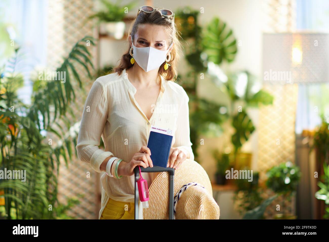 Reise während der Coronavirus-Pandemie. Trendige Reisende Frau mit covid Pass, medizinische Maske, Flugticket, Trolley-Tasche, Desinfektionsmittel und Sonnenbrille in Stockfoto