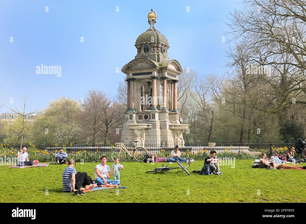 Junge Leute, die im Frühling im Sarphatipark auf Gras relaxen, mit Monument Samuel Sarphati im Hintergrund, De Pijp, Amsterdam, Niederlande, Europa Stockfoto