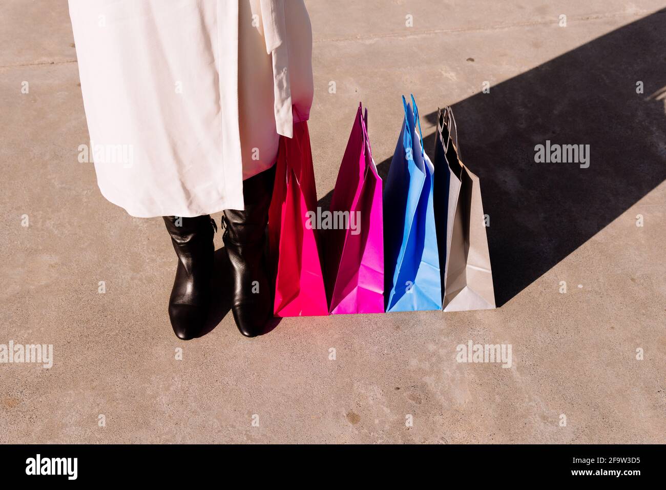 Frauenfüße in Stiefeln und weißem Sommerkleid oder Frühling neben bunten Einkaufstaschen. Einkaufskonzept Stockfoto