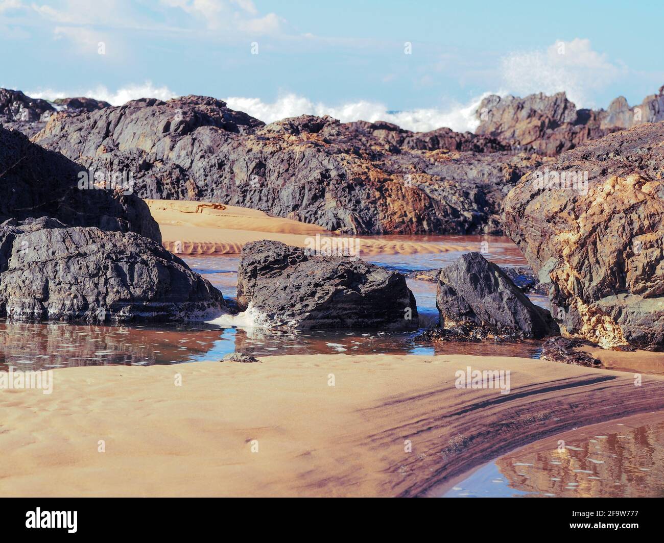Abstrakte Strandszene an der Küste, wunderschöne warme rote Brauntöne mit weitläufigen Linien, natürliche Muster im Sand, Felsenbecken mit krachenden Meereswellen Stockfoto