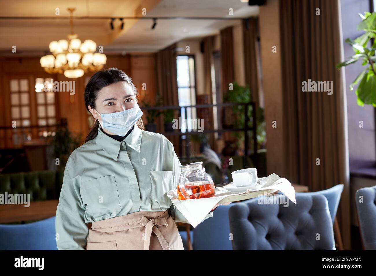 Lächelnde Kellnerin in Maske und Schürze stehend mit Teekannen Tablett im Café und Blick auf die Kamera Stockfoto