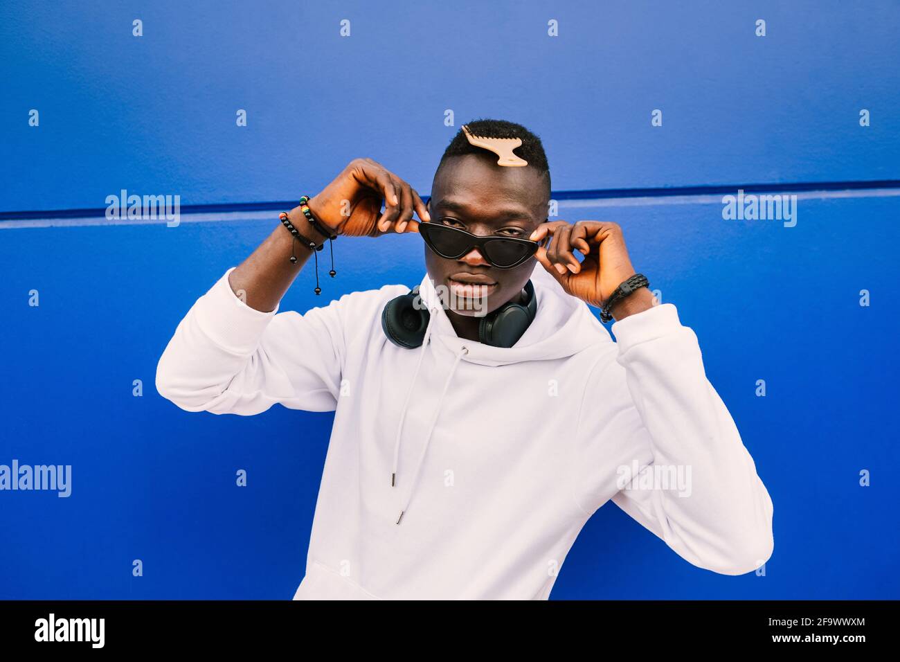 Ein junger afroamerikanischer schwarzer Mann, der ein weißes Sweatshirt, eine Sonnenbrille, einen Kamm und Kopfhörer an einer blauen Wand trägt und voller Zuversicht über die Brille blickt Stockfoto