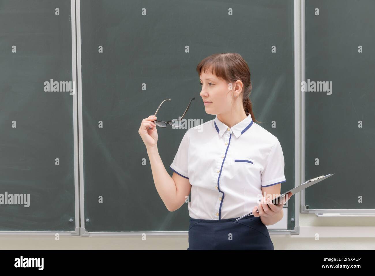 Schullehrerin junge hübsche Frau in einer weißen Bluse mit einem Ordner und einer Brille in den Händen im Klassenzimmer auf dem Hintergrund der Tafel. portr Stockfoto