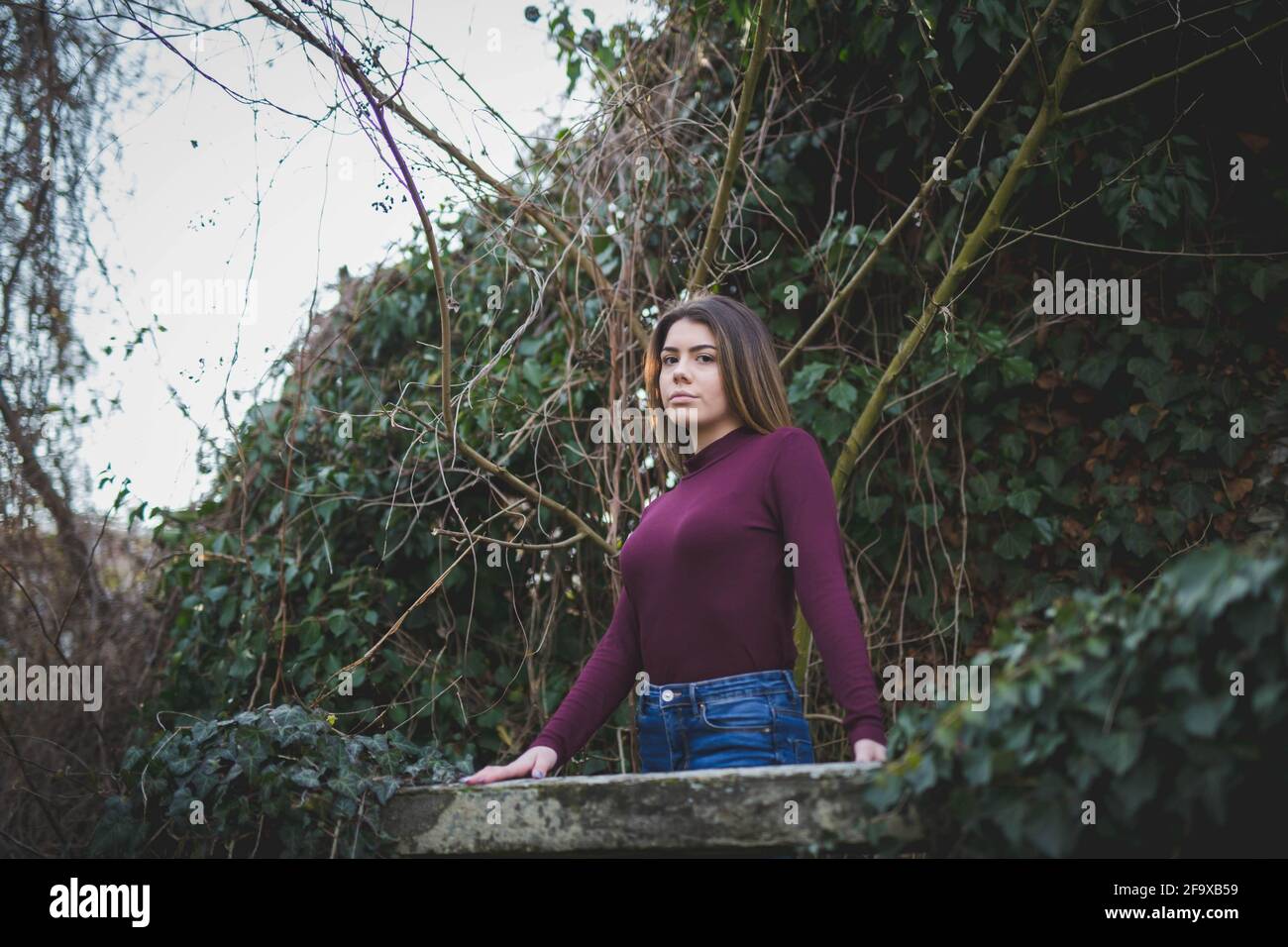 Eine hübsche junge Frau in einem lila Pullover mit Stehkragen Im Freien vor dichtem Grün posieren Stockfoto