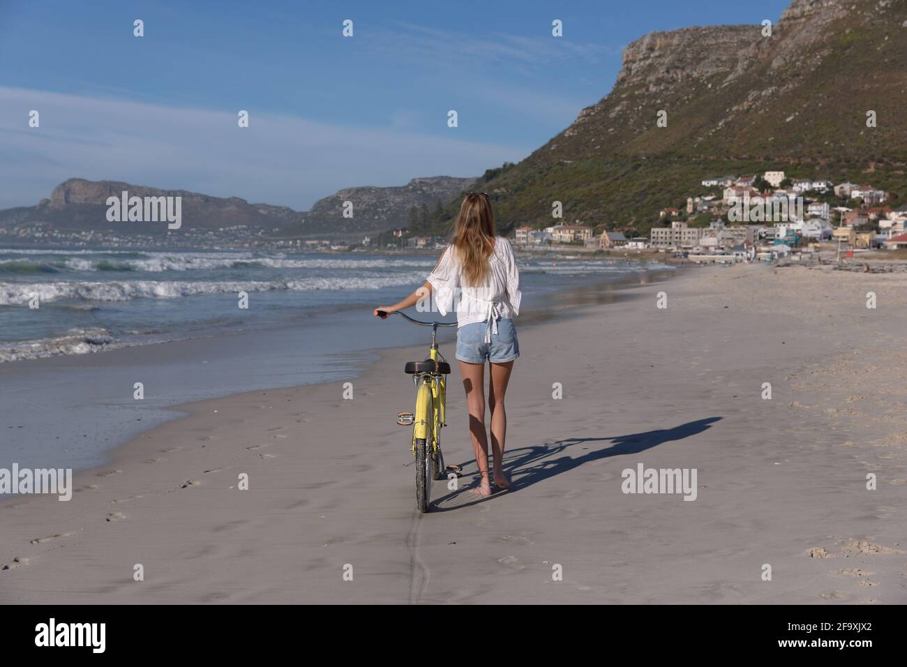 Rückansicht der kaukasischen Frau in weißem Oberteil und Shorts Wandern mit dem Fahrrad am Strand Stockfoto