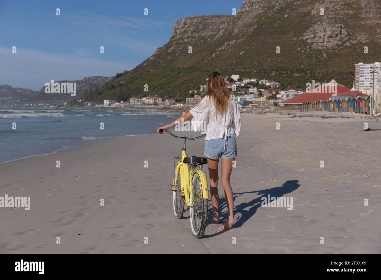 Rückansicht der kaukasischen Frau in weißem Oberteil und Shorts Wandern mit dem Fahrrad am Strand Stockfoto