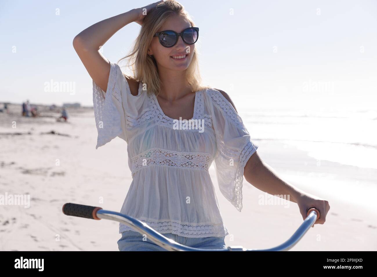 Lächelnde kaukasische Frau mit Sonnenbrille, weißem Oberteil und Shorts, die am Strand auf dem Fahrrad sitzt Stockfoto