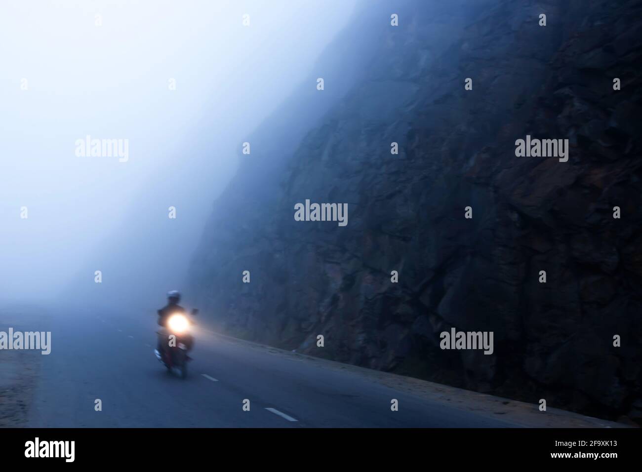 Ein nicht identifizierter Mann, der in der Dämmerung auf der dunklen nebligen Bergstraße Motorrad fährt, eine mystische Asphaltstraße auf einem Pass mit Lichtspur vom Scheinwerfer. Stockfoto