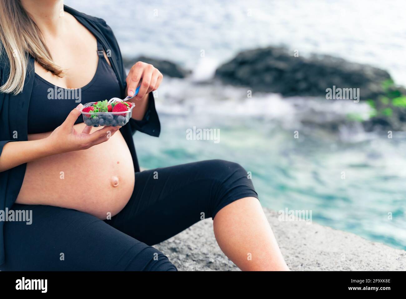 Eine Schwangere sitzt am Meer und isst frischen Obstsalat, Nahaufnahme. Gesunde Ernährung und Liebe für den Foten Konzept. Stockfoto