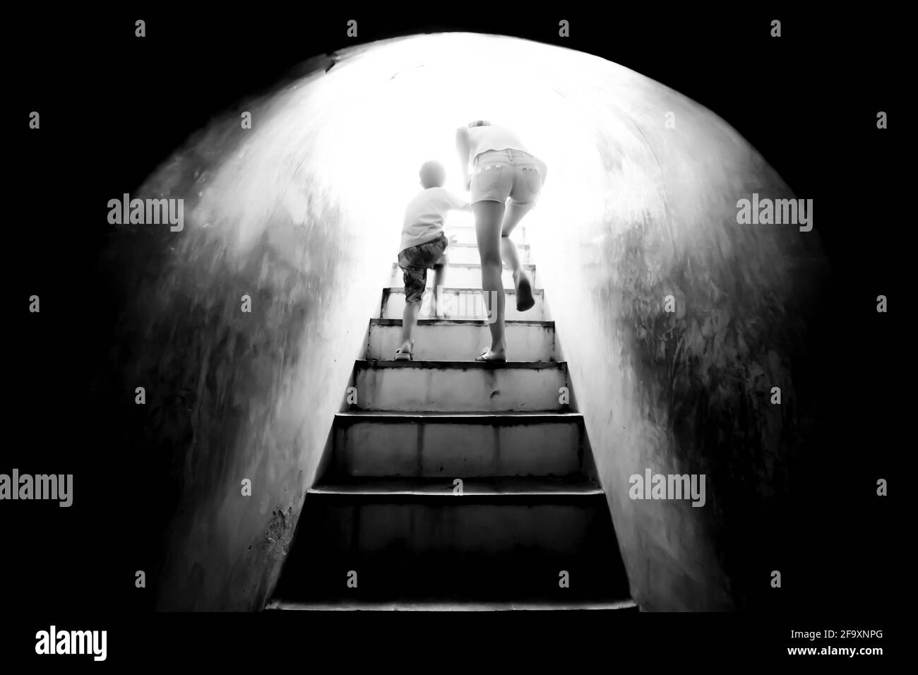 Bewegungsunschärfe. Mutter hält die Hand ihres Sohnes, der die Treppe hinauf zu hellem Sonnenlicht am Ende des dunklen Tunnels geht. Monochrom. Freiheitskonzept. Stockfoto