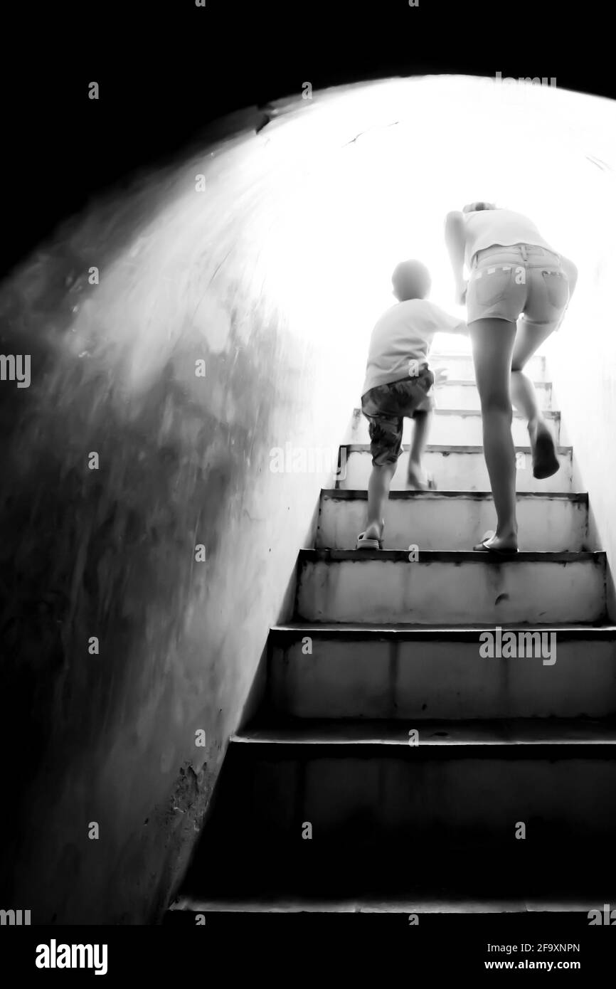 Bewegungsunschärfe. Mutter hält die Hand ihres Sohnes, der die Treppe hinauf zu hellem Sonnenlicht am Ende des dunklen Tunnels geht. Monochrom. Freiheitskonzept. Stockfoto