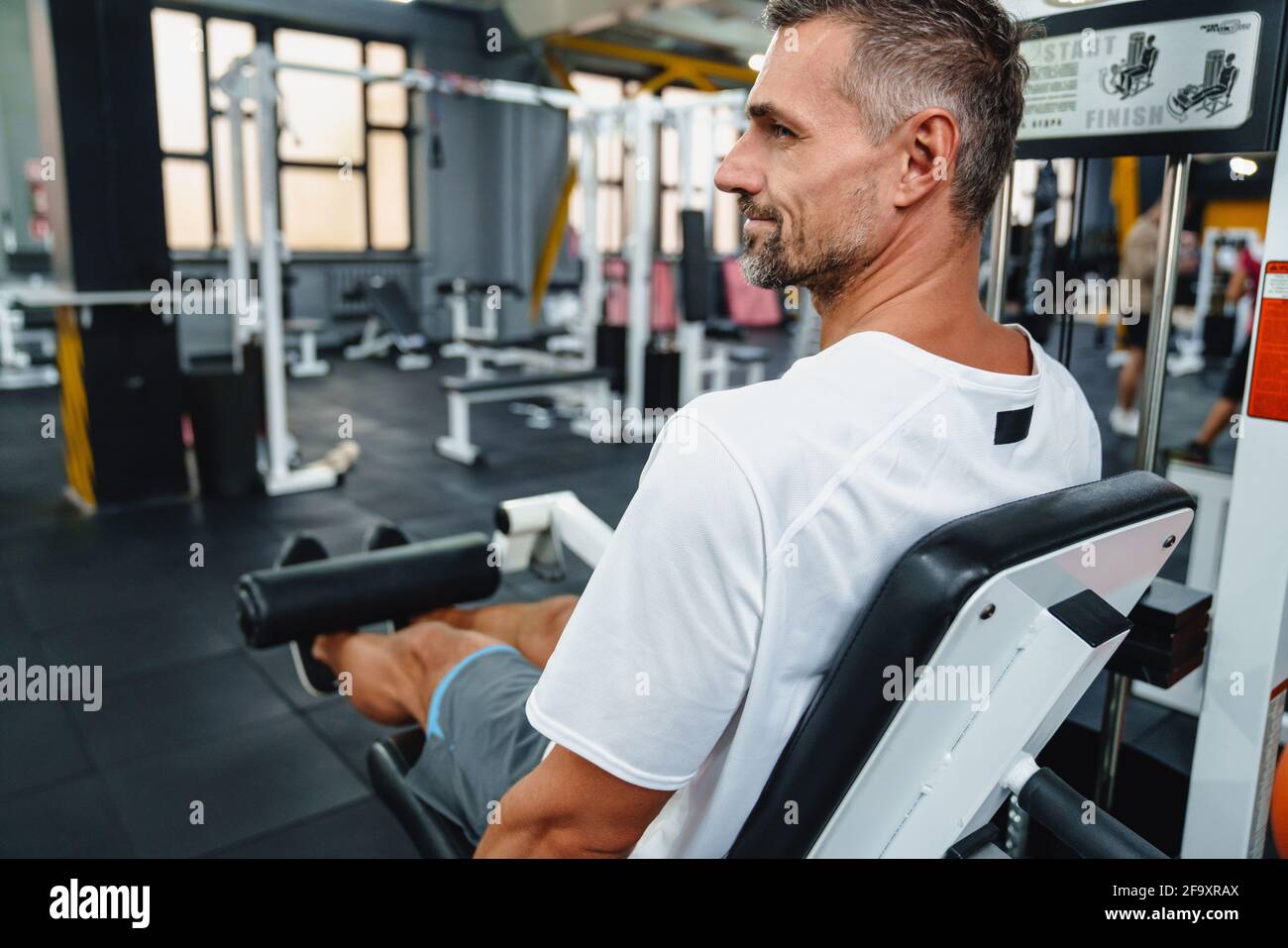 Attraktiver, selbstbewusster, gesunder Sportmann mit sitzender Beincurl-Maschine im Fitnessstudio, Seitenansicht Stockfoto