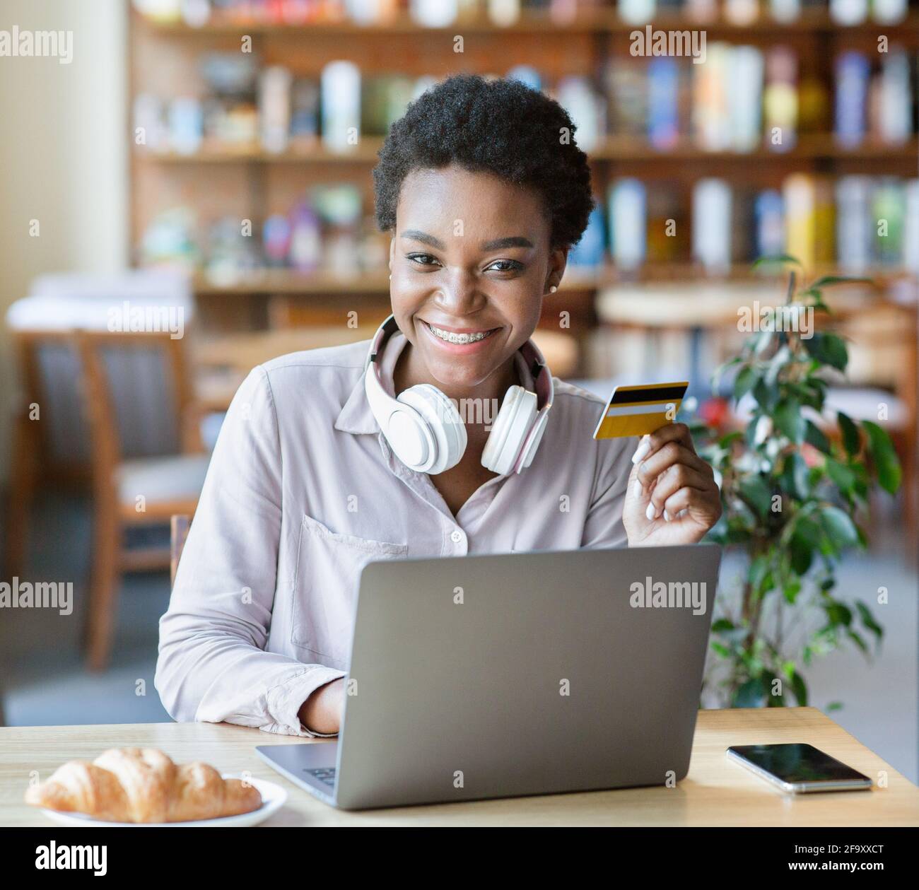 Porträt einer glücklichen jungen schwarzen Frau mit Kopfhörer und Kredit Karte mit Laptop für Online-Shopping im Café Stockfoto