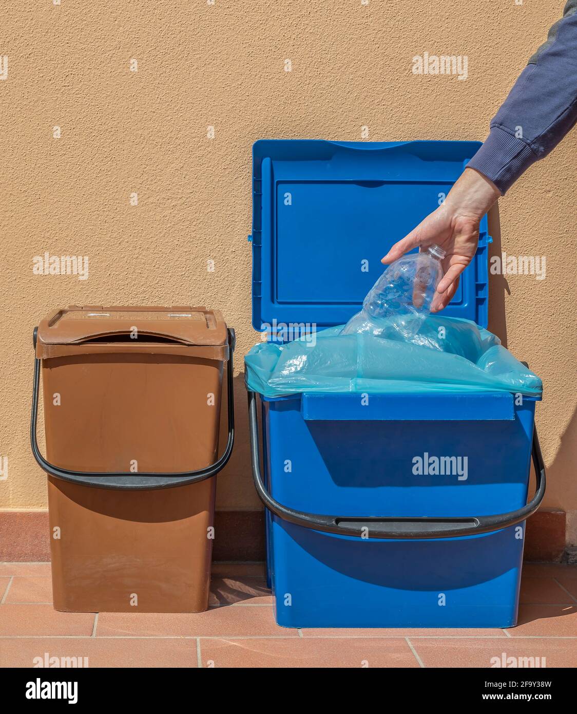 Eine Hand steckt eine leere Plastikflasche in einen blauen Recyclingbehälter, daneben befindet sich ein weiterer brauner Eimer Stockfoto