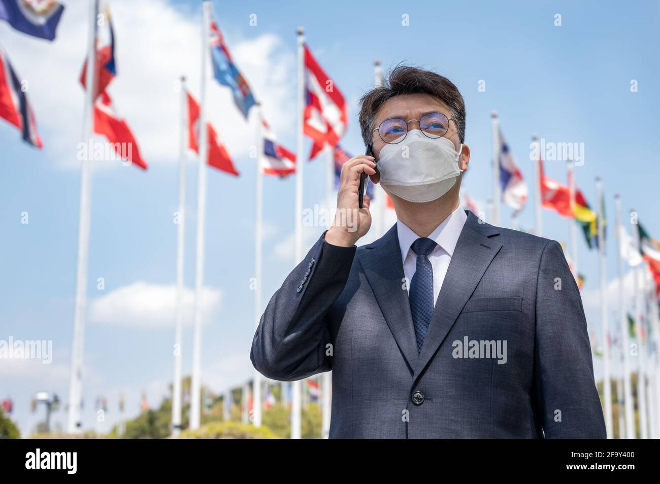 Ein asiatischer Geschäftsmann mittleren Alters trägt eine Maske und benutzt ein Smartphone unter verschiedenen Nationalflaggen, die im Wind flattern. Stockfoto