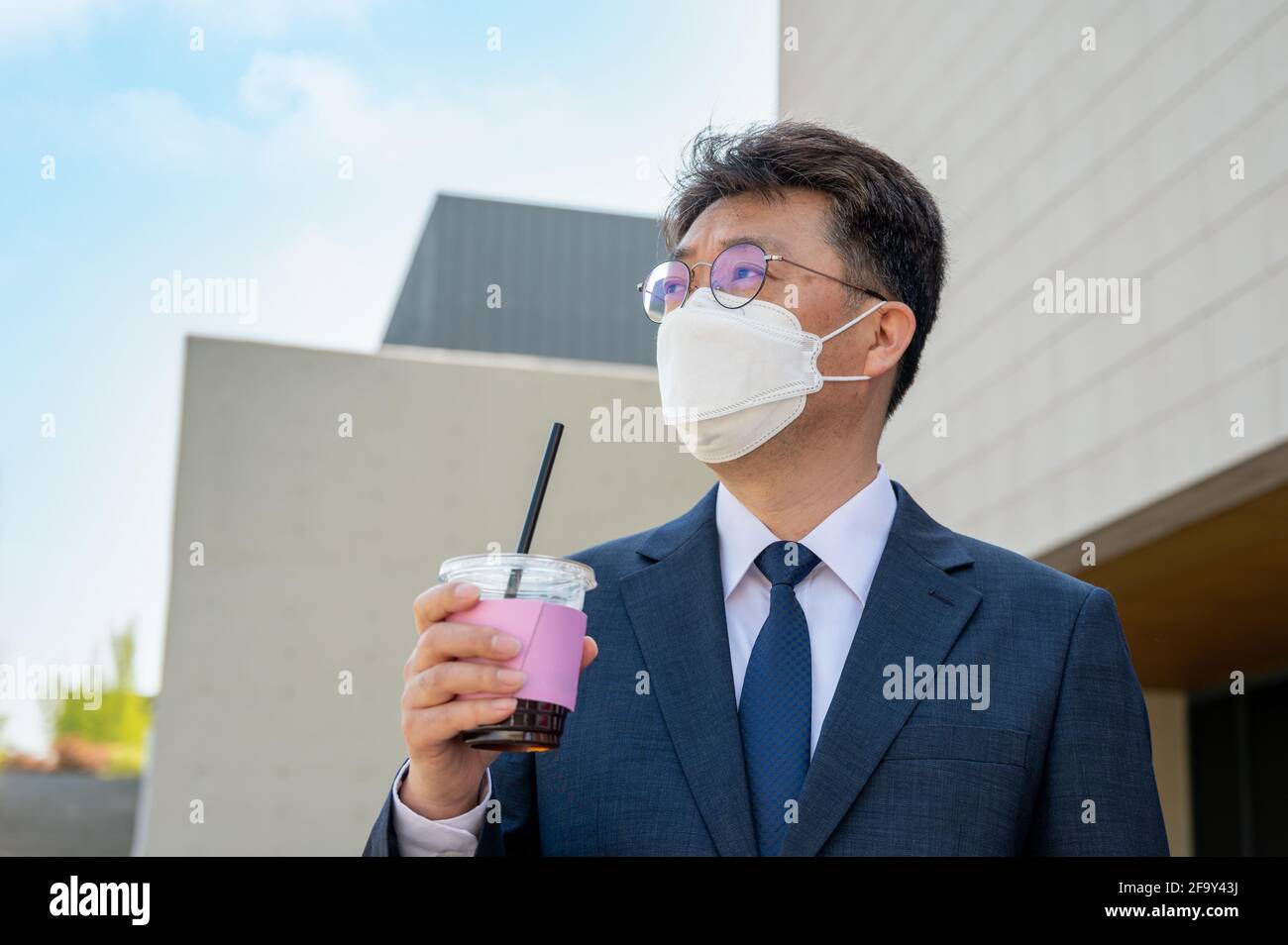 Ein asiatischer Geschäftsmann mittleren Alters in der Stadt, der eine Maske trägt und eine Kaffeetasse hält. Stockfoto