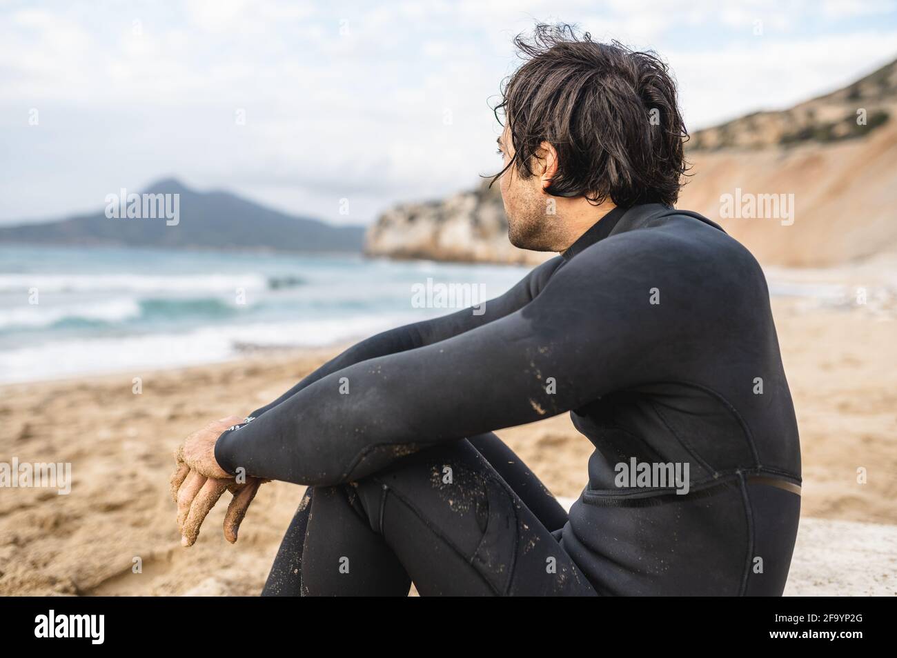 Rückansicht junger Mann mit Tauchanzug sitzt auf dem Sand und blickt auf das Meer. Nasser Mann, der sich nach dem Schwimmen ausruhte. Stockfoto