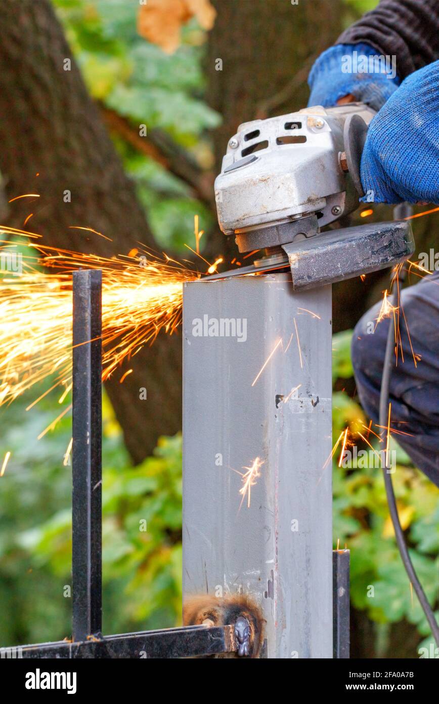 Die Hände des Arbeiters schneiden und waagerecht die Metallpfosten mit einem Winkelschleifer, heiße Funken fliegen während des Metallschneidens heraus. Stockfoto