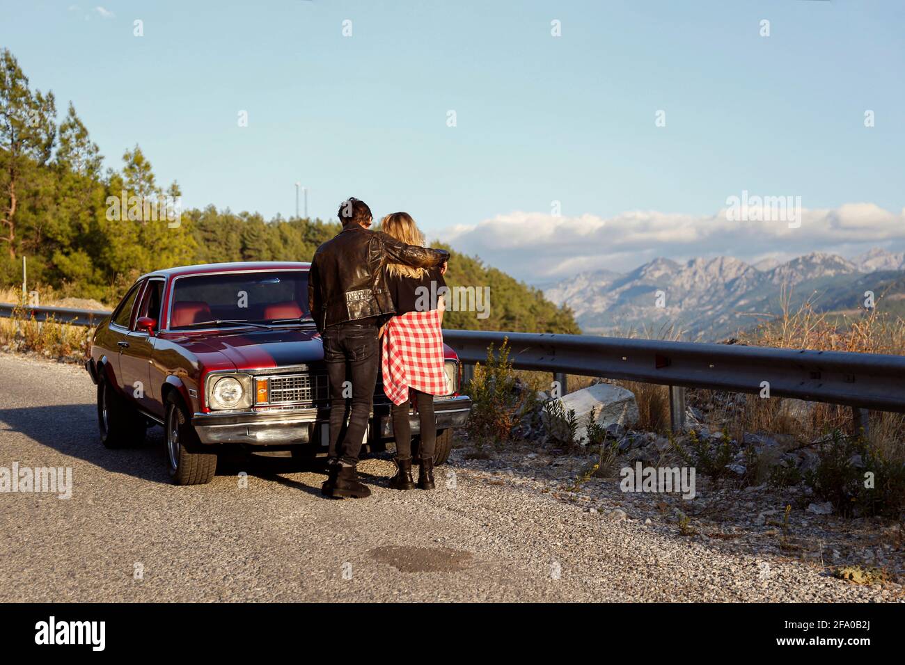 Rückansicht eines Paares, das lässige Kleidung im Rock-Stil trägt, die sich umarmt In der Nähe ihres alten roten Muscle Car auf der Seite geparkt Von der kurvenreichen Straße mit schönen Stockfoto