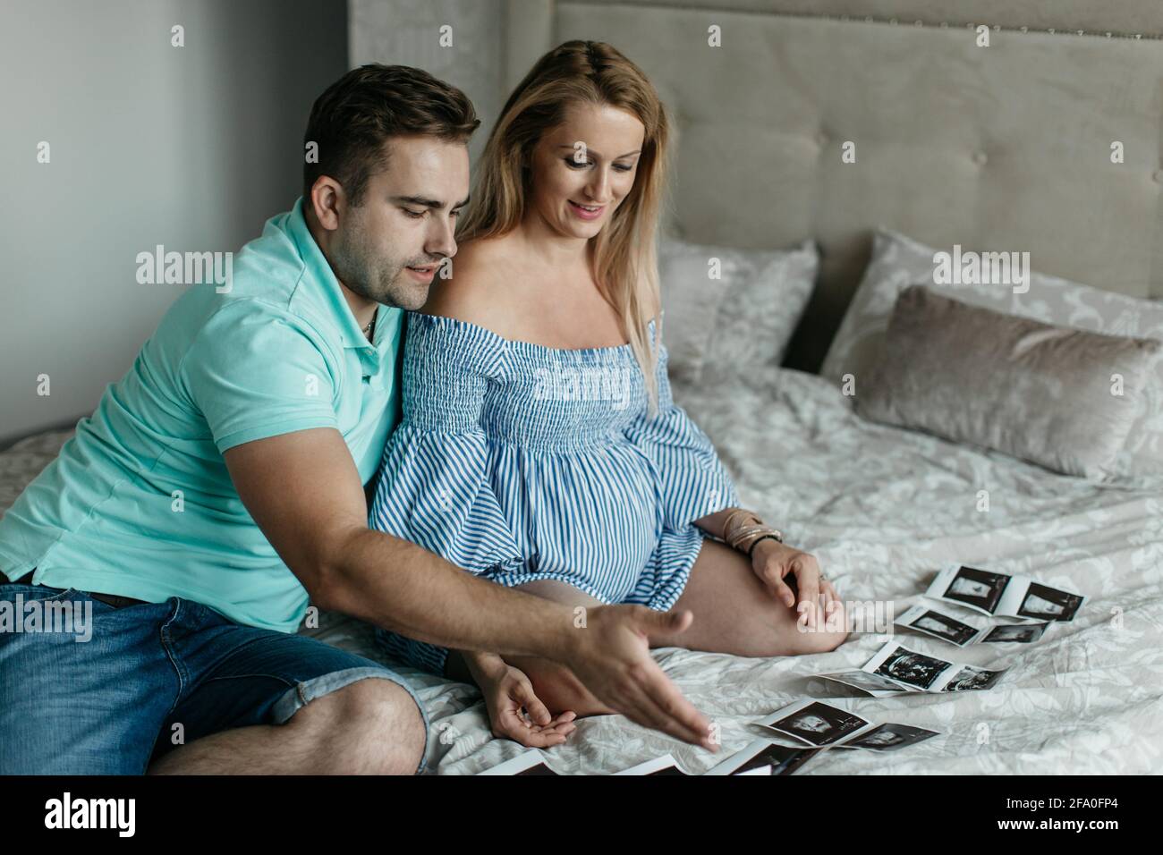 Ein Porträt eines glücklichen Paares, das einen Ultraschall ihres Babys ansieht. Eine lächelnde Schwangerin, die mit ihrem Partner auf einem Bett sitzt und Fotos ansieht. Stockfoto