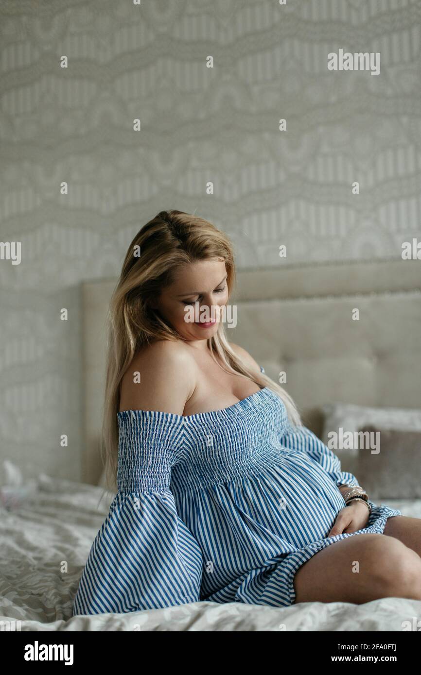 Eine glückliche, schwanger Frau berührt ihren Bauch. Eine fröhliche, werdende Frau, die zu Hause auf einem Bett sitzt und ihre Beule anschaut. Stockfoto