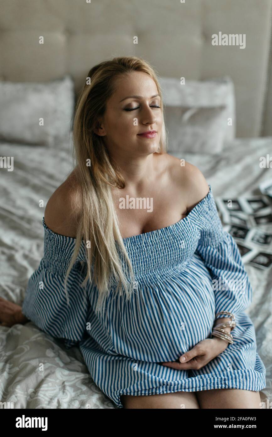 Eine glückliche, schwanger Frau berührt ihren Bauch und träumt davon. Ein Porträt einer wartenden Frau, die zu Hause auf einem Bett sitzt und meditiert. Stockfoto