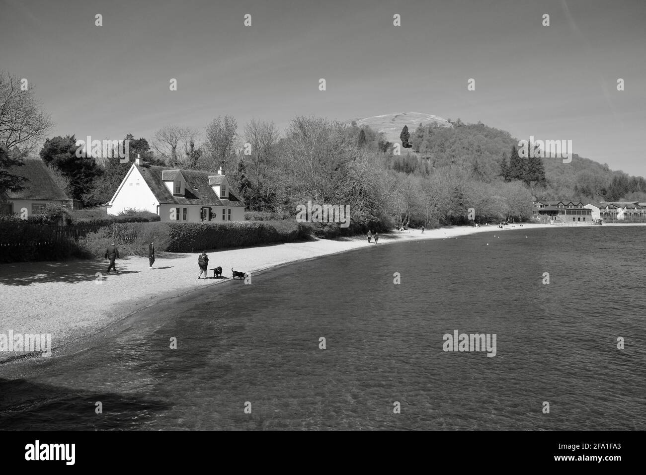 Schwarzweißes landschaftliches Szenario von Menschen, die am Luss-Strand spazieren, mit traditionellen Steinhäusern an den Ufern von Loch Lomond, Schottland, UK Stockfoto
