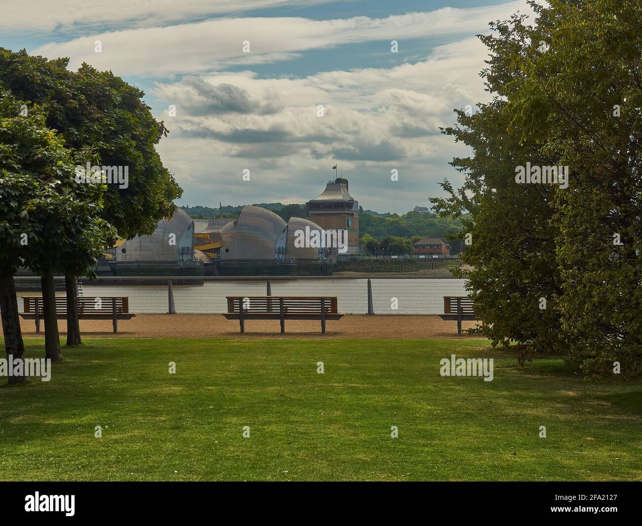 Die metallische, fremde Form der Thames Barrier, von einer Allee aus Bäumen auf der Nordseite des Flusses, mit einem sommerlichen Himmel aus blauen und flauschigen Wolken. Stockfoto