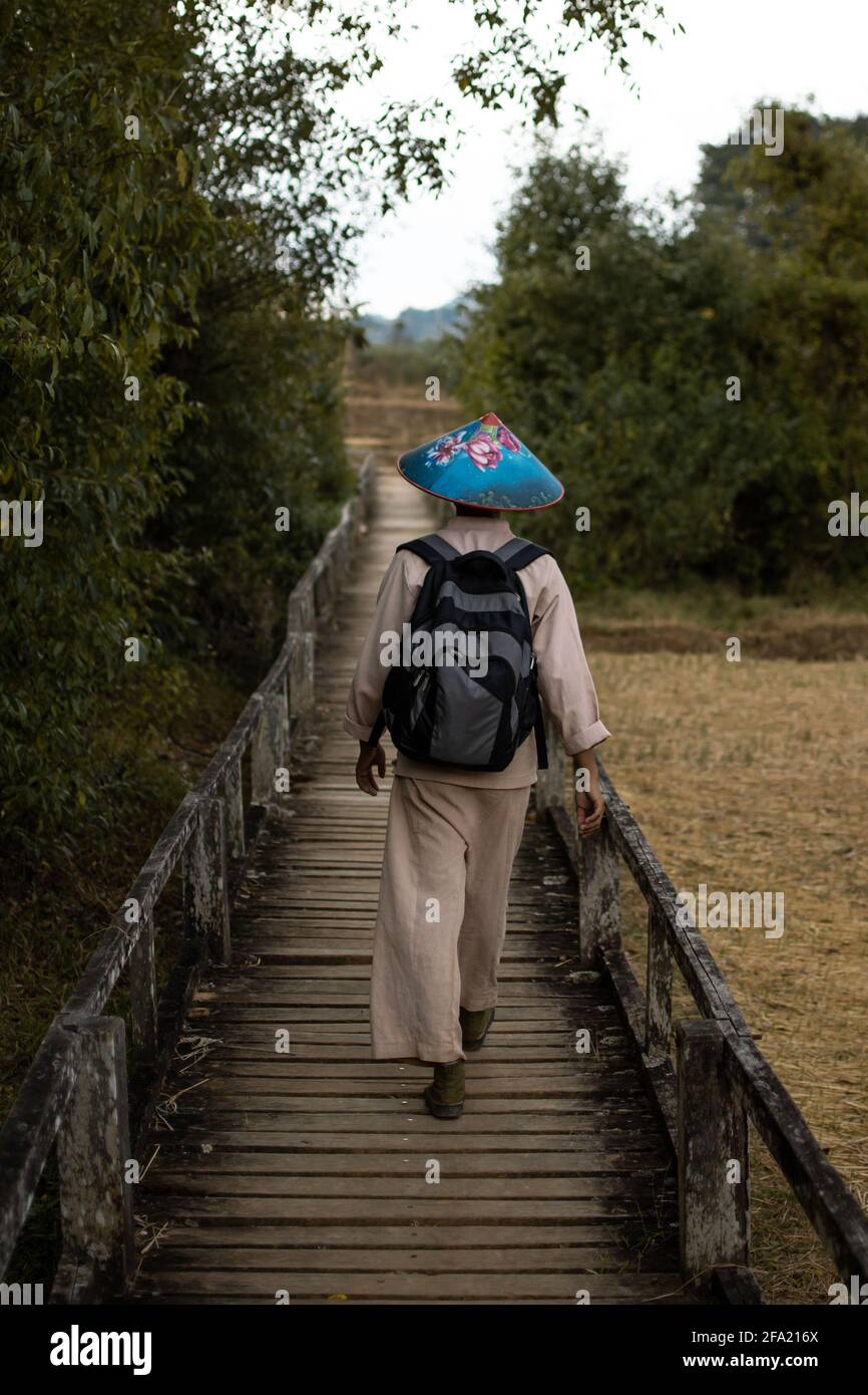 Burmesischer Mann in traditioneller Kleidung und einem farbenfrohen blauen Hut wandern auf einem Gehweg vorbei an Reisfeldern von Kalaw zum Inle Lake, Shan State, Myanmar Stockfoto