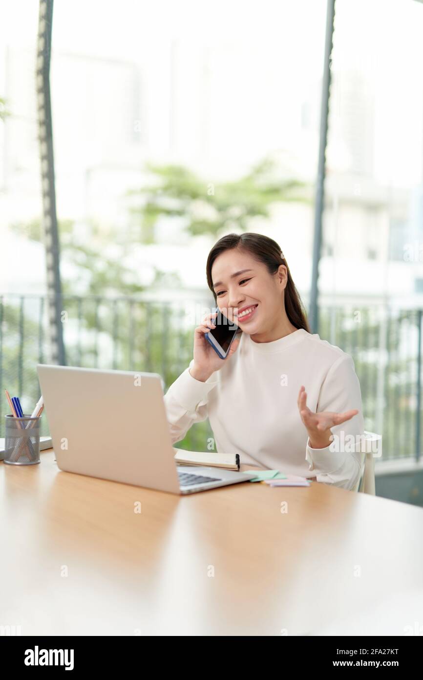 Digitaler Nomade. Junge Unternehmerin, die mit dem Handy spricht, auf den Laptop schaut und lächelt, glückliche Frau, die im Café oder von zu Hause aus arbeitet. Stockfoto