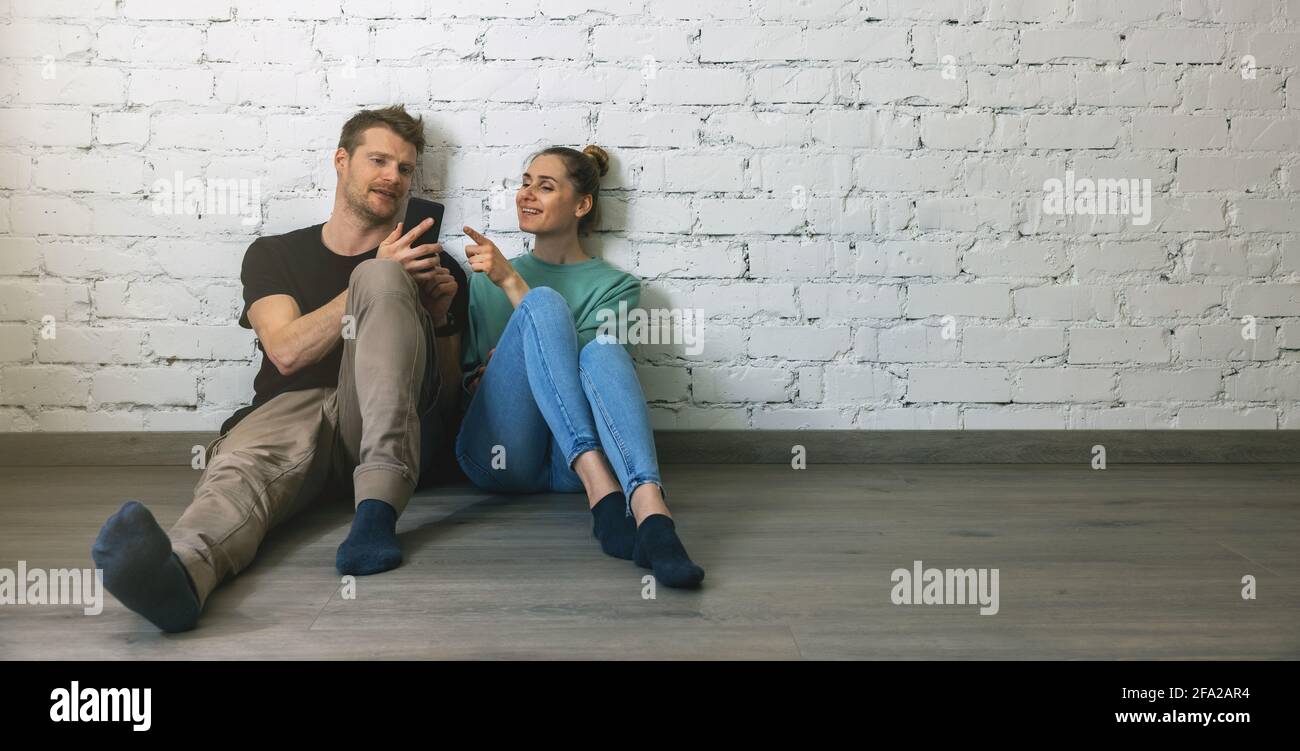 Paar sitzt auf dem Boden an der Wand in leeren Raum und mit dem Telefon, um Möbel online zu wählen. Umzug in ein neues Zuhause Konzept Stockfoto