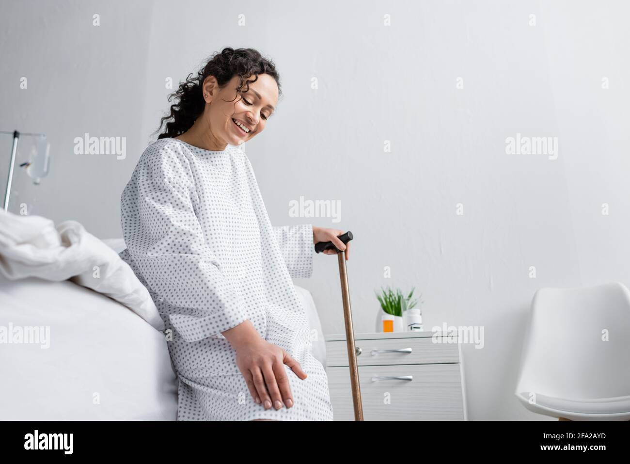 Positive afroamerikanische Frau berührt Knie, während sie auf dem Bett sitzt Mit Gehstock Stockfoto
