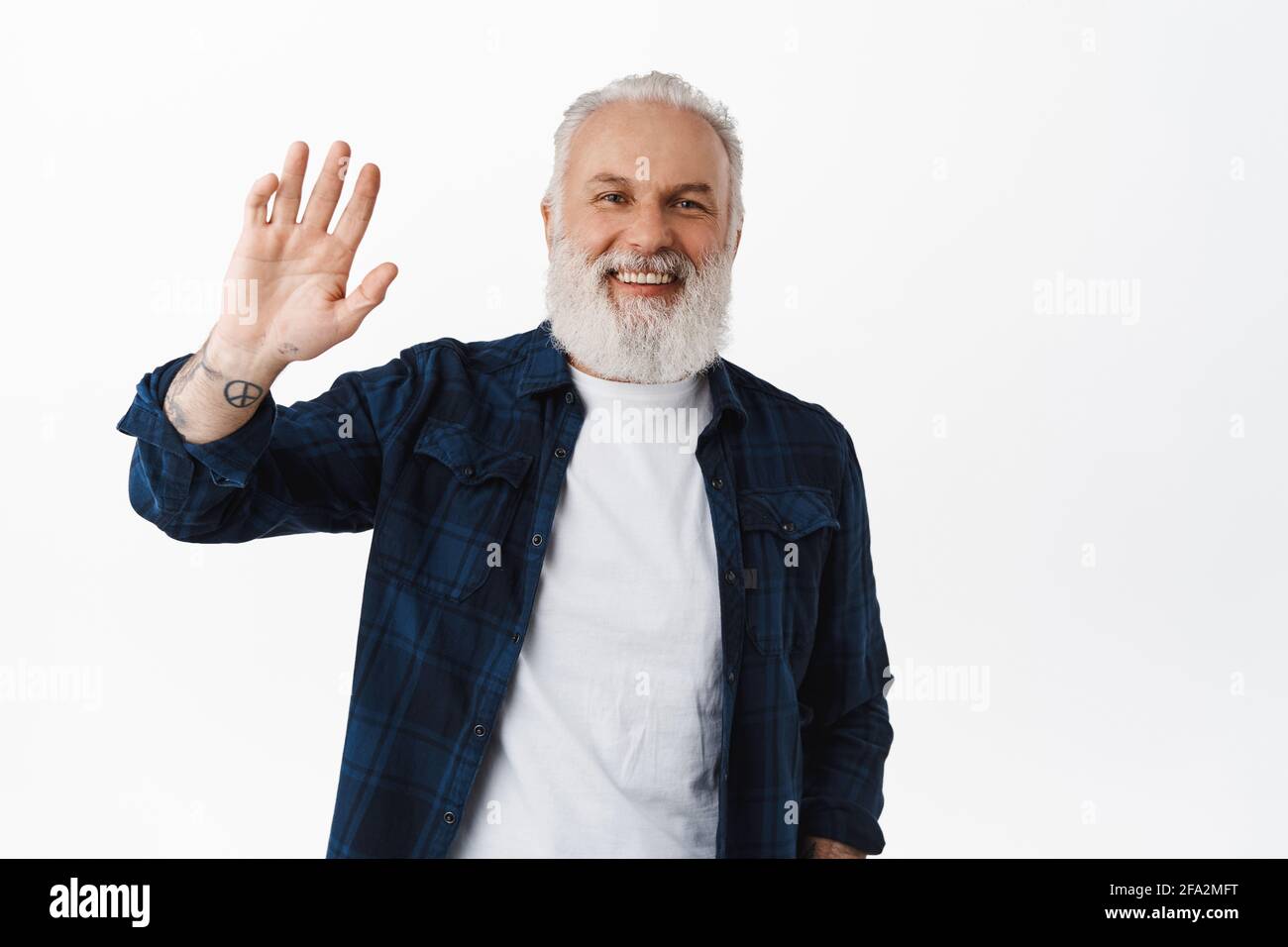 Freundlicher alter Mann winkt Hand, um Hallo. zu sagen Lächelnder Senior mit Tattoos winkt als Gruß, macht hallo Geste, steht glücklich gegen Weiß Stockfoto