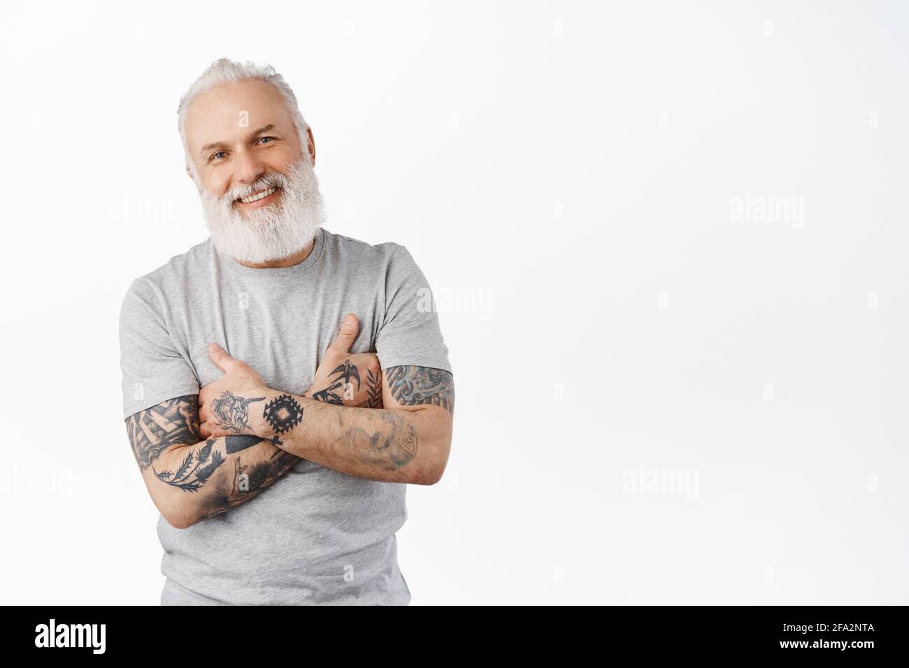 Stilvoller reifer Mann mit Tattoos und langen Bart-Kreuzarmen auf der Brust, lächelnd und offen auf die Kamera schauend. Authentischer, sorgloser älterer Mann im Stehen Stockfoto