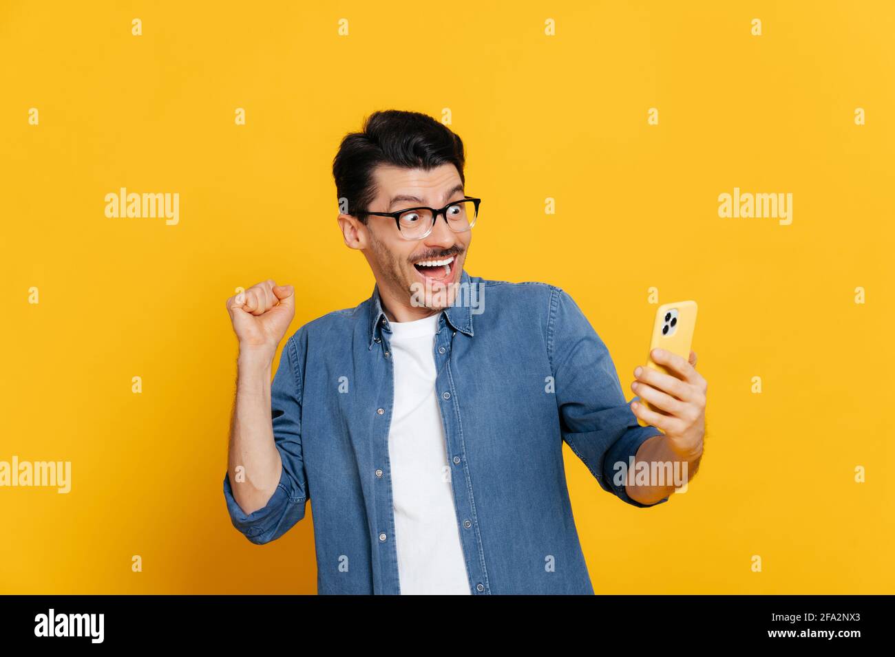 Stauned fröhlich aufgeregt stilvolle kaukasischen Kerl hält Smartphone, erhalten unerwartete Nachrichten, gewinnen, steht auf isoliertem orangefarbenen Hintergrund, fröhlichen Gesichtsausdruck, toothy Lächeln, gestikulieren mit Faust Stockfoto