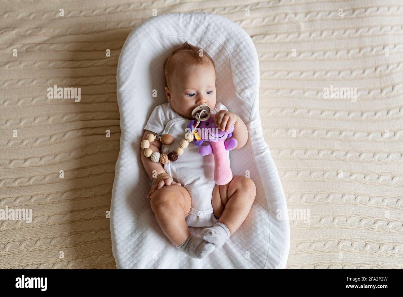 Niedliches blondes Baby, drei Monate alt, das zu Hause im Kokon liegt. Kind hält ausgestopftes Spielzeug, saugt Nippel. Stockfoto