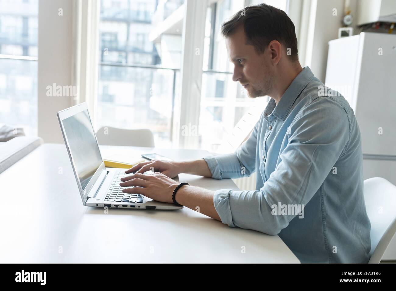 Seitenansicht eines konzentrierten jungen Geschäftsmannes, der auf der Tastatur tippt, eines intelligenten Mannes, der im eleganten Casual-Hemd E-Mails beantwortet und online auf dem Laptop von zu Hause aus rempt arbeitet Stockfoto