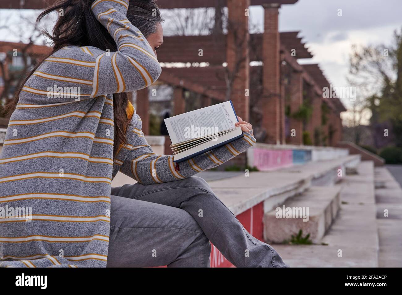 Frau, die ein Buch liest und dabei die Haare von ihrem Gesicht streicht, sitzt auf einem Steinschritt, der von der Seite betrachtet wird Stockfoto