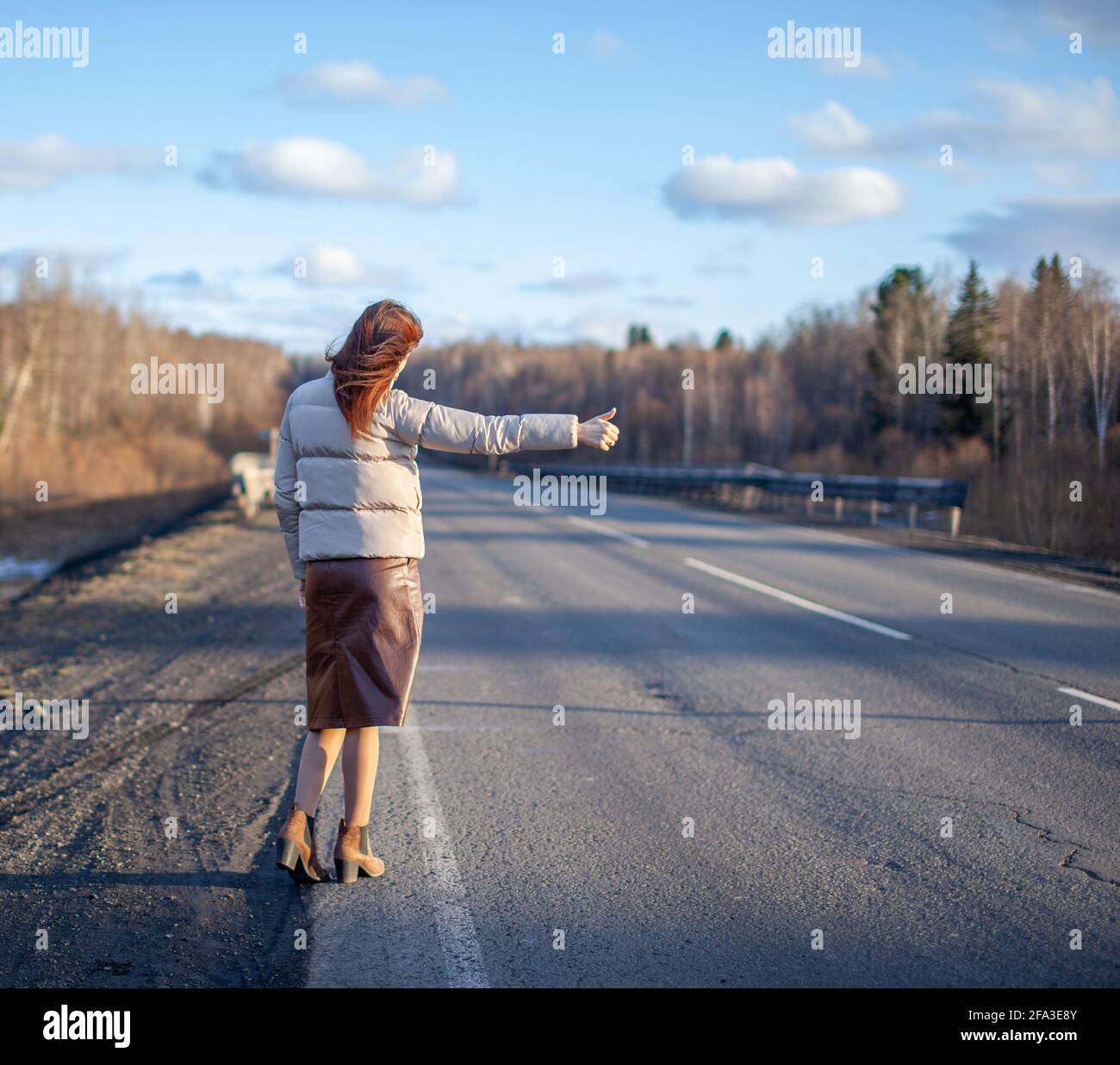 Das Mädchen hält das Auto mit der Hand auf der Autobahn an. Stilvolle Frau auf der Straße hält das Auto auf eine Reise gehen. Eine Straße mitten im Wald. Stockfoto