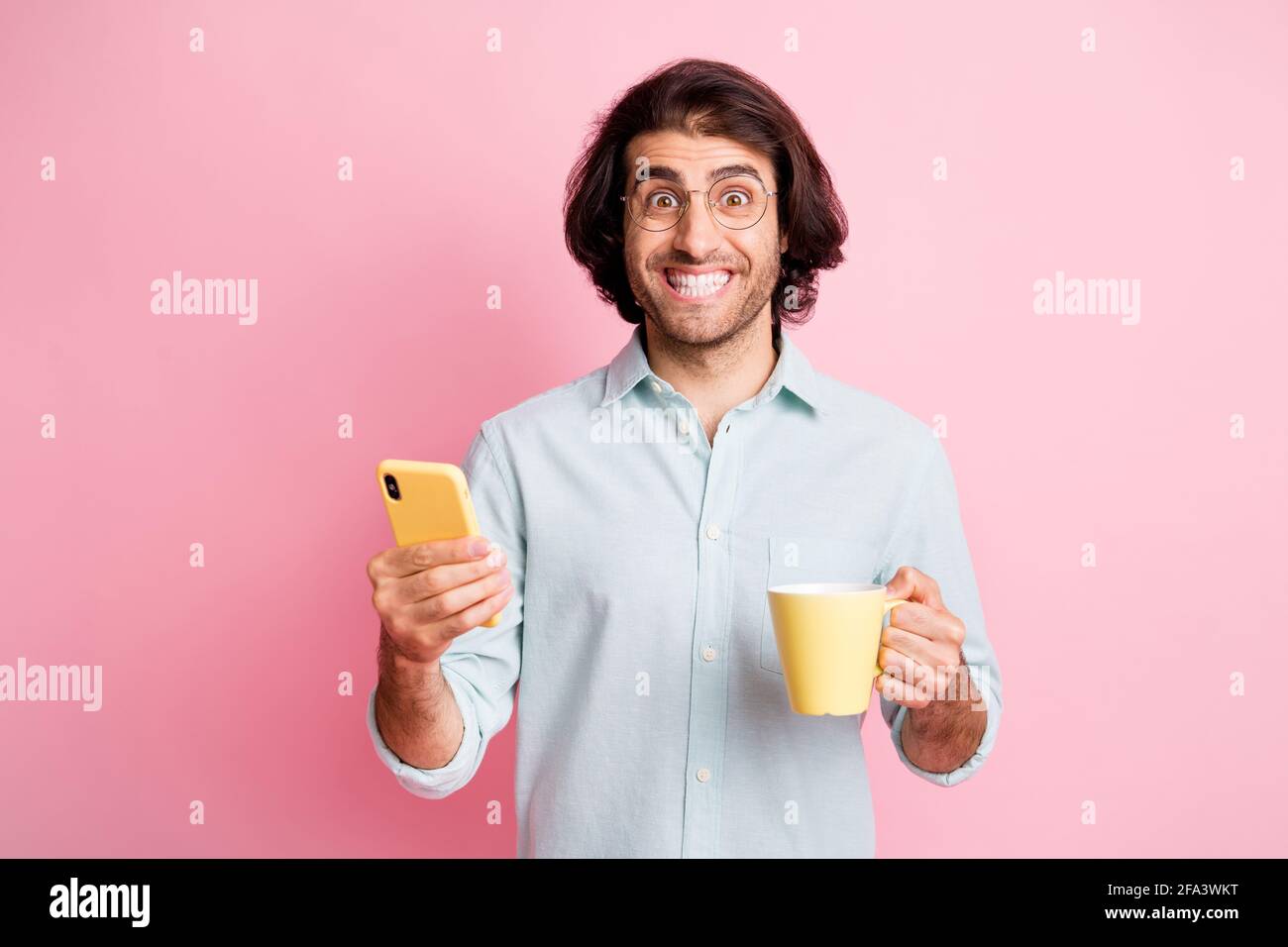 Foto von gut aussehenden fröhlich braunen Haar Kerl halten Tasse Telefon Tragen Sie Brillen blaues Hemd isoliert auf pastellrosa Hintergrund Stockfoto