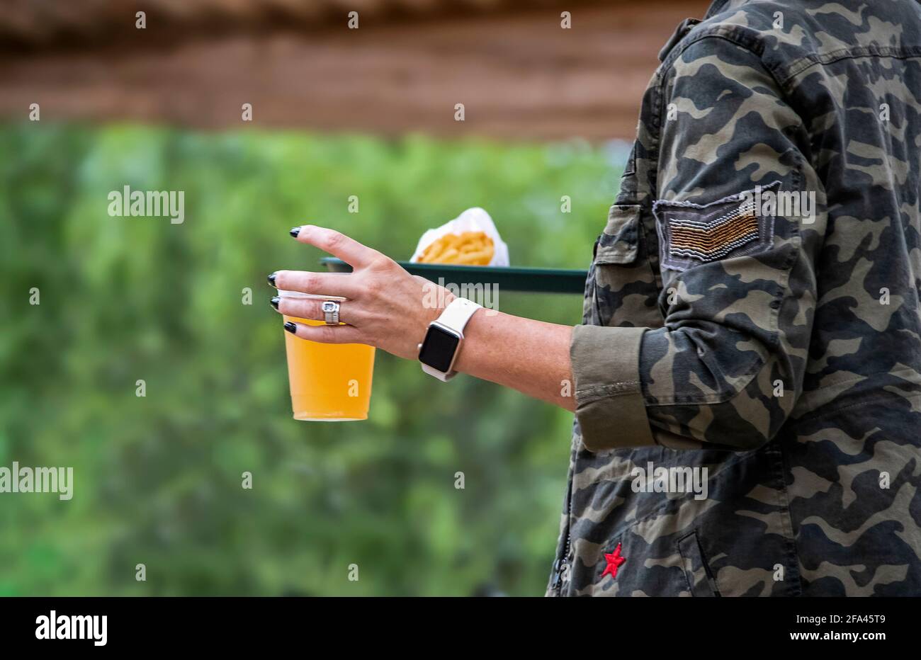 Frau mit schwarzen Fingernägeln und Camojacke trägt Tablett mit Essen und Bier auf einer Seite des Rahmens mit halten Bokeh-Bäume und Außenschutzdach in BA Stockfoto