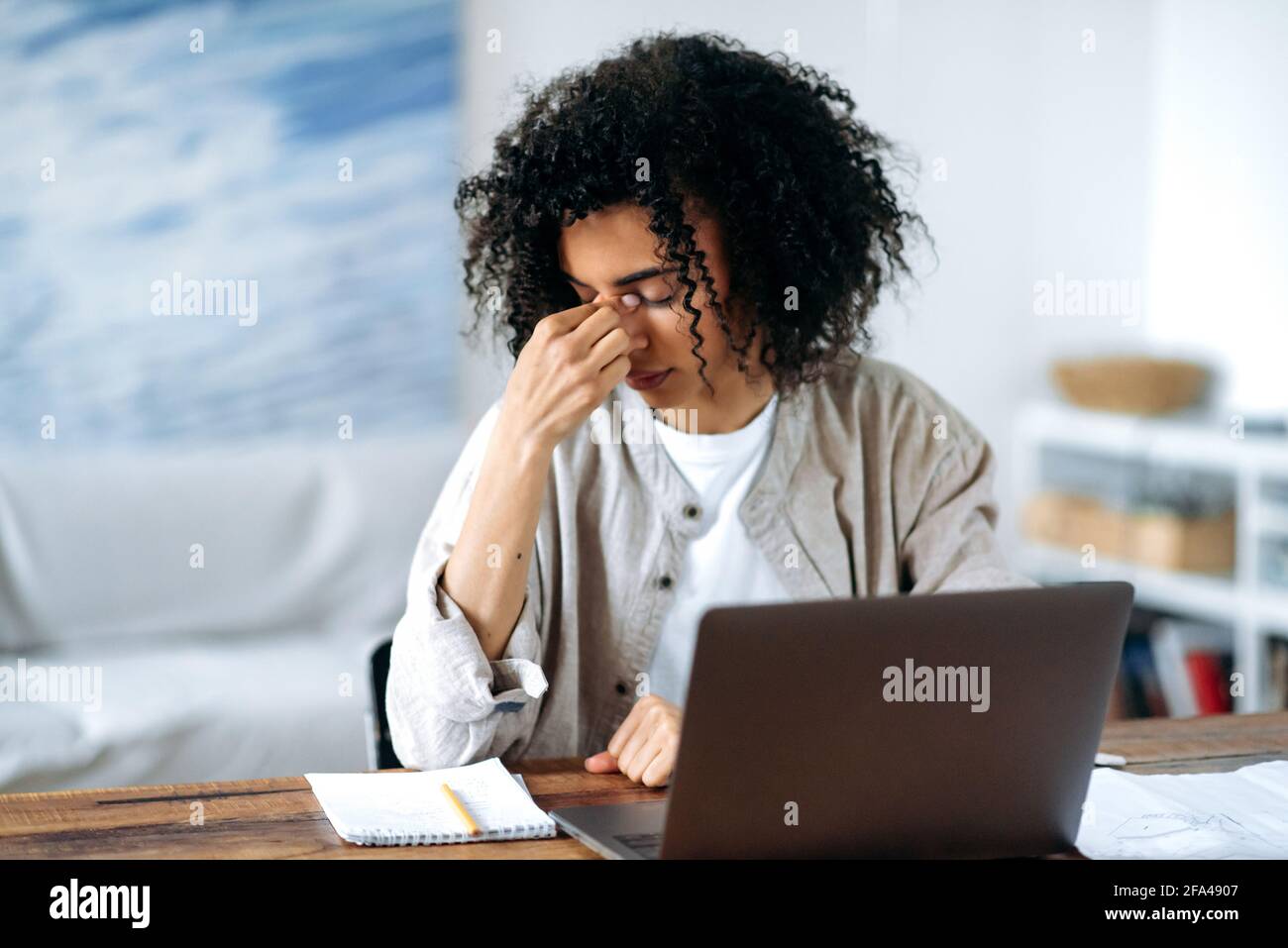 Müde frustrierte überarbeitete afroamerikanische junge Frau in Freizeitkleidung, Managerin, Freiberuflerin oder Studentin, die im Wohnzimmer am Laptop sitzt, Stress, Kopfschmerzen, Reib und ihre Augen schließt Stockfoto