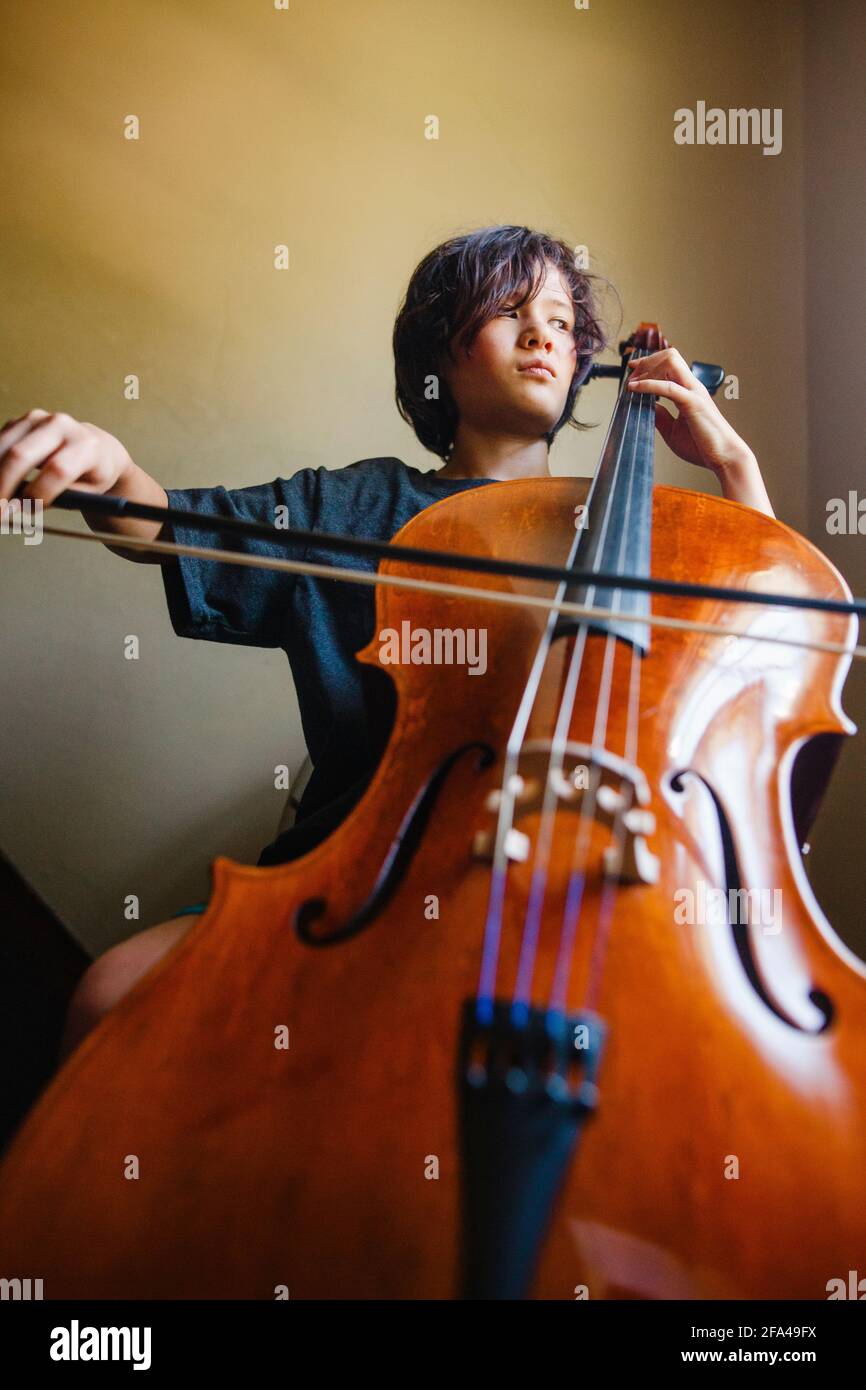 Ein Junge mit ernsthafteren Ausdrücken spielt beim Schauen Cello Aus dem Fenster Stockfoto